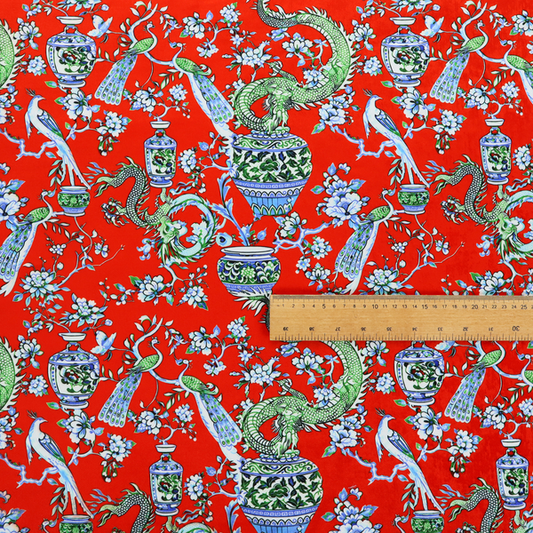 Vibrant red Chinoiseries fabric featuring an intricate pattern of green dragons, elegant peacocks, blue-and-white porcelain vases, and blossoming floral branches.