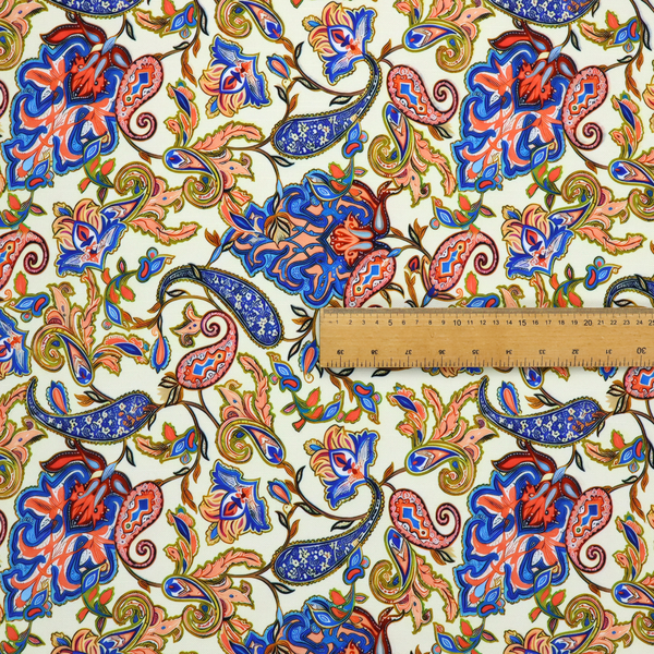 Cream-coloured fabric with an ornate repeating paisley pattern showcasing royal blue tear-drop motifs, vibrant coral and orange florals, and flowing leafy vines.