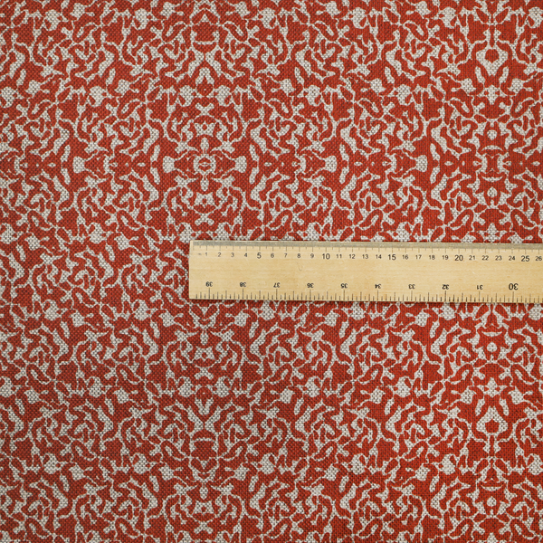 A decorative textile featuring a symmetrical, kaleidoscopic pattern in red and beige. The intricate, interlocking organic shapes create a dense, filigree-like design with a slightly weathered or distressed look. The fabric has a visible, fine-textured weave that gives the pattern a soft, matte appearance.