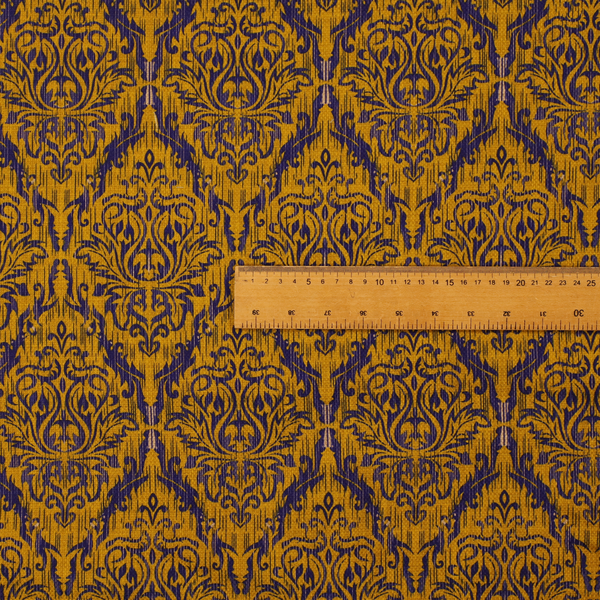 This fabric features a royal blue damask pattern layered over a deep mustard gold background. The design has an ikat-inspired finish, with vertical, feathered edges that give the ornate fleur-de-lis motifs a slightly blurred, textured appearance.