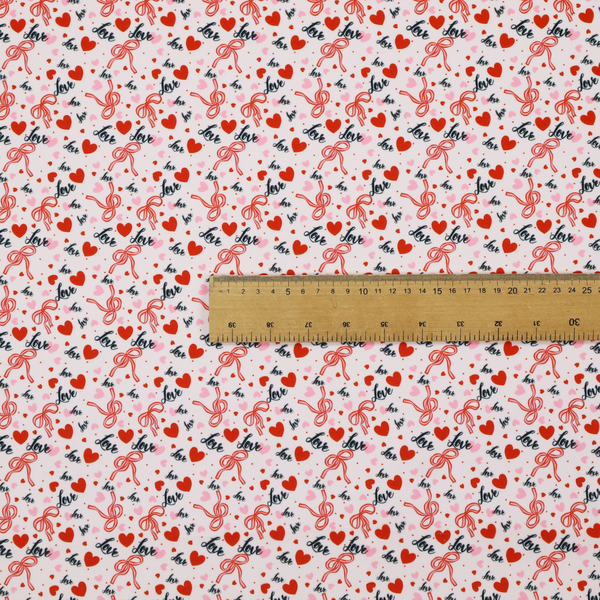 This light pink fabric features a dense, repeating Valentine’s Day pattern with a vintage, romantic aesthetic. The design is composed of small red hearts and larger pink hearts, interspersed with delicate red ribbon bows and the word "love" written in a dark, flowing cursive script. Tiny red dots are scattered throughout the background, creating a textured, confetti-like effect across the entire print.