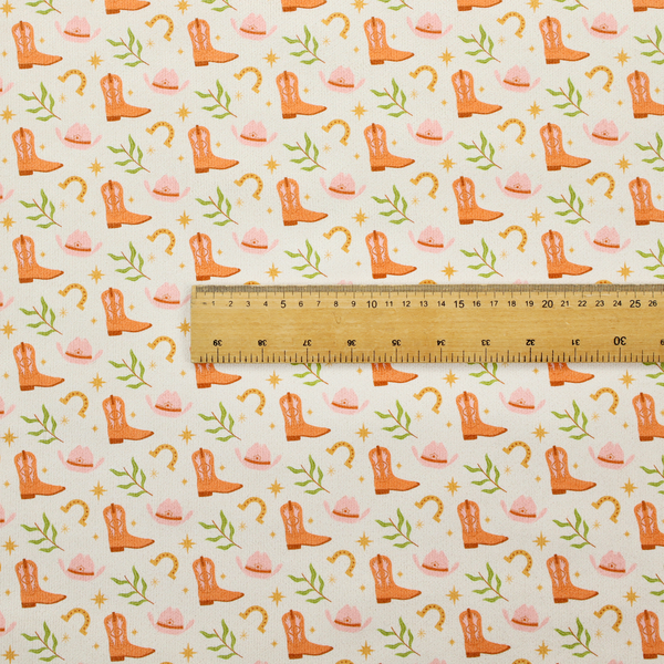 A repeating pattern of cowboy-themed illustrations on a cream-coloured fabric backdrop. The design features light orange cowboy boots, pale pink cowboy hats, golden horseshoes, small green leafy sprigs, and delicate four-pointed stars scattered throughout.