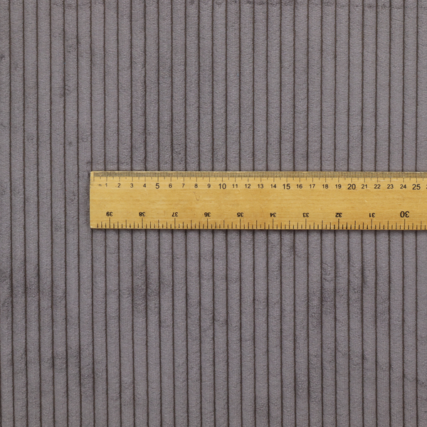 A corduroy fabric with a visible, vertical ribbed texture. A wooden ruler is placed across the fabric to provide a scale, showing approximately 4 to 5 ribs per centimetre.