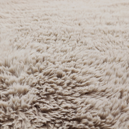 A taupe or light greyish-brown, high-pile faux fur fabric. It features a dense, plush texture with soft, tousled fibres that create a cosy, shag-like appearance.