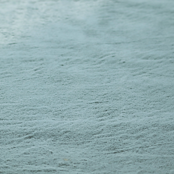 A plush faux fur fabric in a muted dusty teal or slate blue shade. The material has a dense, short-pile texture that creates a soft, uniform surface with a gentle matte sheen. The fibres are tightly packed, showing subtle tonal variations and light shadows that give the fabric a velvety depth.