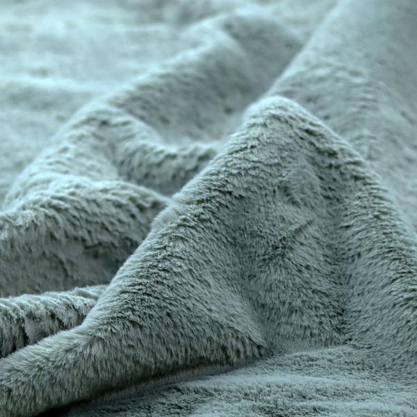 A plush faux fur fabric in a muted dusty teal or slate blue shade. The material has a dense, short-pile texture that creates a soft, uniform surface with a gentle matte sheen. The fibres are tightly packed, showing subtle tonal variations and light shadows that give the fabric a velvety depth.