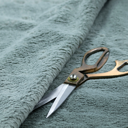 A plush faux fur fabric in a muted dusty teal or slate blue shade. The material has a dense, short-pile texture that creates a soft, uniform surface with a gentle matte sheen. The fibres are tightly packed, showing subtle tonal variations and light shadows that give the fabric a velvety depth.