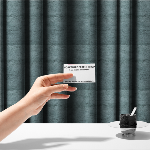 The fabric used to create these made-to-measure curtains is a plush faux fur fabric in a muted dusty teal or slate blue shade. The material has a dense, short-pile texture that creates a soft, uniform surface with a gentle matte sheen. The fibres are tightly packed, showing subtle tonal variations and light shadows that give the fabric a velvety depth.