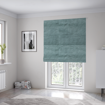 The fabric used to create these Roman blinds is a plush faux fur fabric in a muted dusty teal or slate blue shade. The material has a dense, short-pile texture that creates a soft, uniform surface with a gentle matte sheen. The fibres are tightly packed, showing subtle tonal variations and light shadows that give the fabric a velvety depth.