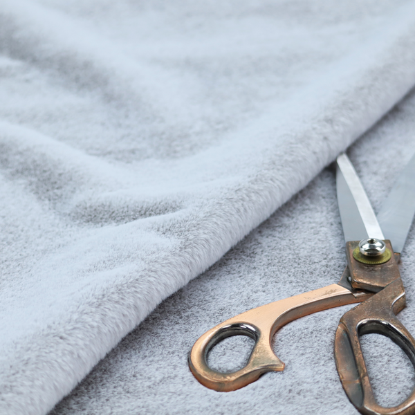 A plush faux fur fabric in a bright silver or icy light grey shade. The material has a dense, short-pile texture that creates a soft, uniform surface with a delicate matte sheen. The cool, pale grey fibres are tightly packed, showing subtle light-and-shadow variations that give the fabric a clean, velvety depth.