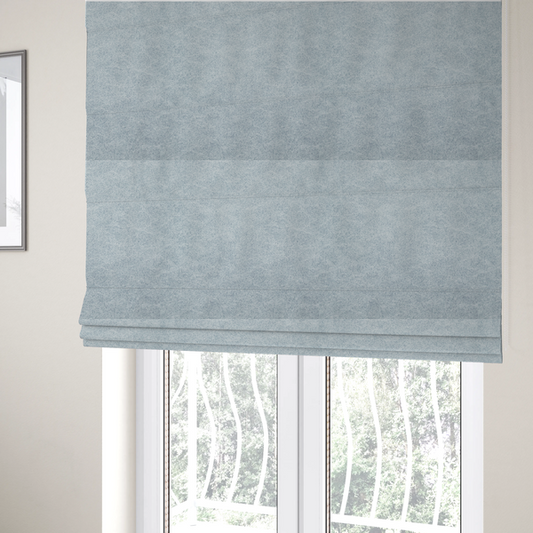 The fabric used to create these Roman blinds is a light sky blue or powder blue fabric with a dense, distressed felt-like texture. The surface features a soft, mottled blend of pale blue and white fibres, creating a subtle weathered effect. It includes the signature irregular, light-coloured "crackle" lines seen throughout this collection, giving it the appearance of weathered microfiber or industrial wool.