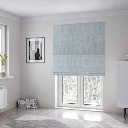 The fabric used to create these Roman blinds is a light sky blue or powder blue fabric with a dense, distressed felt-like texture. The surface features a soft, mottled blend of pale blue and white fibres, creating a subtle weathered effect. It includes the signature irregular, light-coloured "crackle" lines seen throughout this collection, giving it the appearance of weathered microfiber or industrial wool.