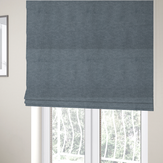 The fabric used to create these Roman blinds is a denim or dusty blue fabric with a dense, distressed felt-like texture. The surface features an intricate mottled blend of slate blue and muted grey fibres. Like the others in this collection, it is defined by organic, light-coloured "crackle" lines and a stippled appearance that mimics the look of weathered suede or high-end industrial wool.