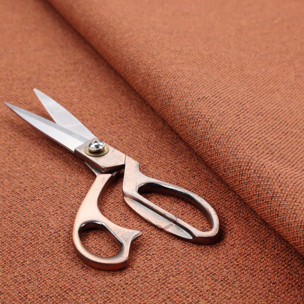 A deep rust or burnt orange textile with a dense diagonal twill weave. The fabric features a rich, multi-tonal blend of saturated orange yarns interlaced with darker brown and subtle flecks of green or gold, giving it a heathered, earthy depth.