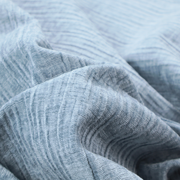 A textured upholstery fabric in a soft powder blue or light sky blue shade. The material features a distinct cross-hatched slub texture with subtle vertical and horizontal ridges, creating a soft, dimensional chenille finish.