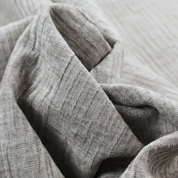 A textured upholstery fabric in a mid-tone grey or pebble shade. The material features a distinct cross-hatched slub texture with prominent vertical and fine horizontal ridges, creating a soft, dimensional chenille finish.