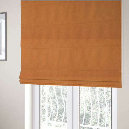 The fabric used to create these Roman blinds is a bright orange or tangerine Merida Suede upholstery fabric. The textile features a highly detailed, dense, and uniform embossed pebbled pattern, giving the soft suede material an intricate, tactile shagreen texture.