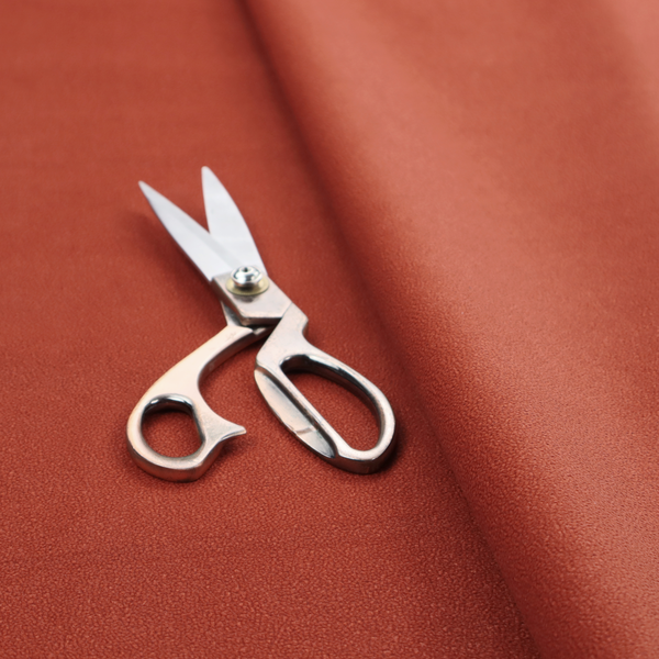 A vibrant brick red or rust Merida Suede upholstery fabric. The textile features a highly detailed, dense, and uniform embossed pebbled pattern, giving the soft suede material an intricate, tactile shagreen texture.