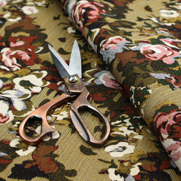 A repeating vintage floral pattern on a warm gold and beige textured woven fabric background. The design features dense clusters of blooming roses in shades of dusty pink, deep red, cream, and muted lavender, surrounded by olive green and dark foliage.   
