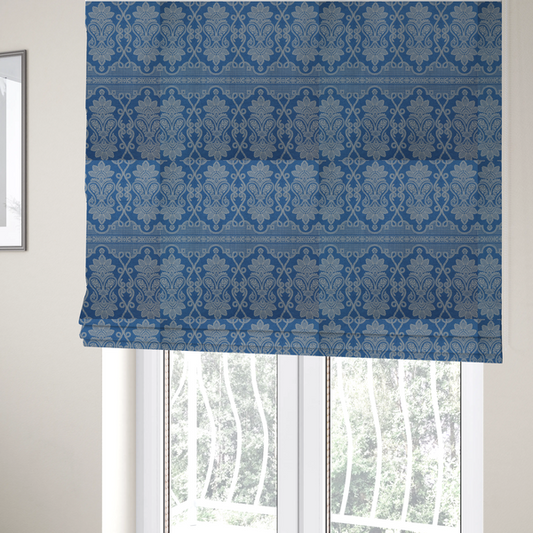 The fabric used to create these Roman blinds is a ornate, traditional damask upholstery fabric on a rich blue background featuring intricate cream-coloured symmetrical floral and paisley motifs divided horizontally by a decorative lattice and scroll border.   