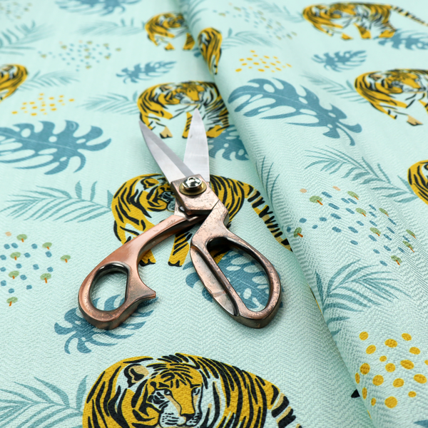 A tropical jungle upholstery fabric on a light mint-blue background featuring a repeating pattern of yellow and black tigers surrounded by teal monstera leaves, palm fronds, and yellow dot clusters.   