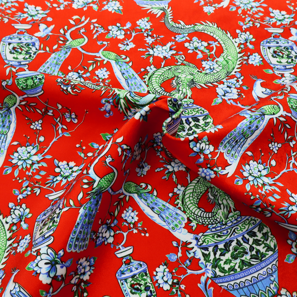 Vibrant red Chinoiseries fabric featuring an intricate pattern of green dragons, elegant peacocks, blue-and-white porcelain vases, and blossoming floral branches.