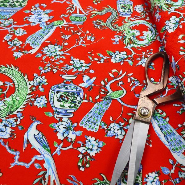 Vibrant red Chinoiseries fabric featuring an intricate pattern of green dragons, elegant peacocks, blue-and-white porcelain vases, and blossoming floral branches.