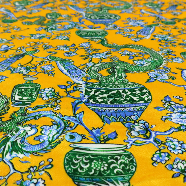 Seamless fabric pattern featuring Chinoiserie-style green dragons, blue and white peacocks, ornate vases, and floral branches against a vibrant mustard yellow background.