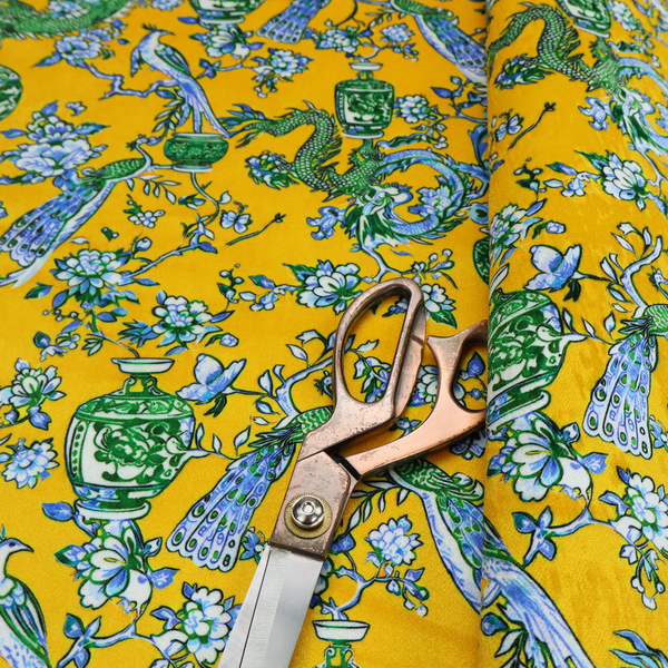 Seamless fabric pattern featuring Chinoiserie-style green dragons, blue and white peacocks, ornate vases, and floral branches against a vibrant mustard yellow background.