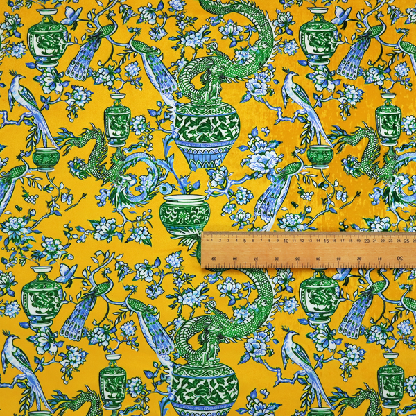 Seamless fabric pattern featuring Chinoiserie-style green dragons, blue and white peacocks, ornate vases, and floral branches against a vibrant mustard yellow background.