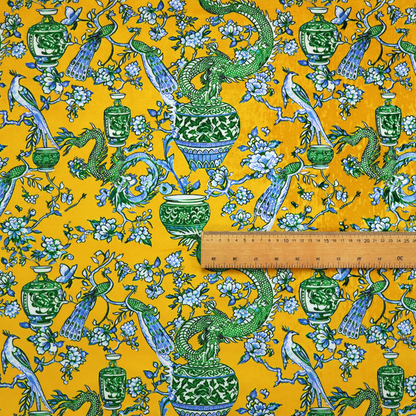 Seamless fabric pattern featuring Chinoiserie-style green dragons, blue and white peacocks, ornate vases, and floral branches against a vibrant mustard yellow background.