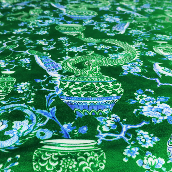 Emerald green fabric featuring a repeating Chinoiserie pattern with light green dragons, blue and white porcelain vases, perched peacocks, and floral branches.   