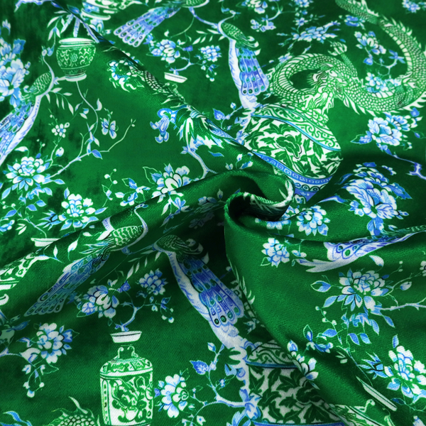Emerald green fabric featuring a repeating Chinoiserie pattern with light green dragons, blue and white porcelain vases, perched peacocks, and floral branches.   
