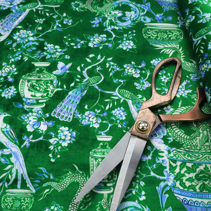 Emerald green fabric featuring a repeating Chinoiserie pattern with light green dragons, blue and white porcelain vases, perched peacocks, and floral branches.   