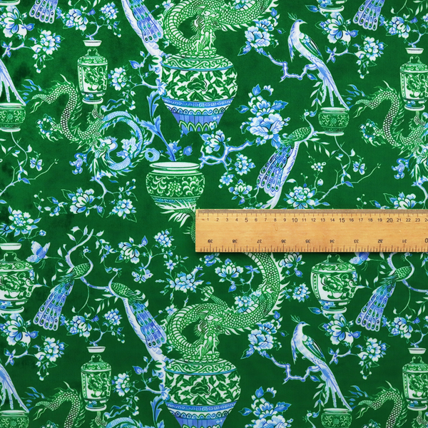 Emerald green fabric featuring a repeating Chinoiserie pattern with light green dragons, blue and white porcelain vases, perched peacocks, and floral branches.   