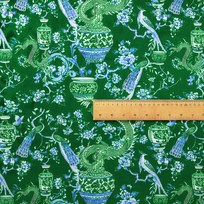 Emerald green fabric featuring a repeating Chinoiserie pattern with light green dragons, blue and white porcelain vases, perched peacocks, and floral branches.   