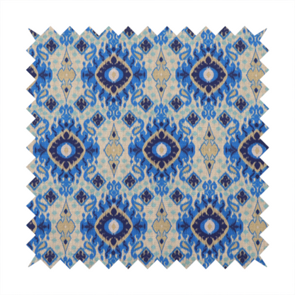 A repeating textile pattern featuring ornate blue Ikat medallions with a feathered, hand-painted appearance. The design includes smaller navy diamonds and sandy beige geometric shapes set against a light oatmeal-coloured fabric texture.