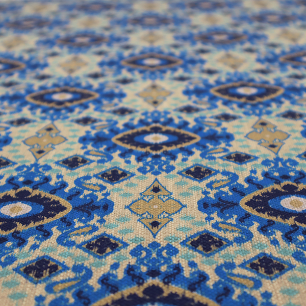 A repeating textile pattern featuring ornate blue Ikat medallions with a feathered, hand-painted appearance. The design includes smaller navy diamonds and sandy beige geometric shapes set against a light oatmeal-coloured fabric texture.