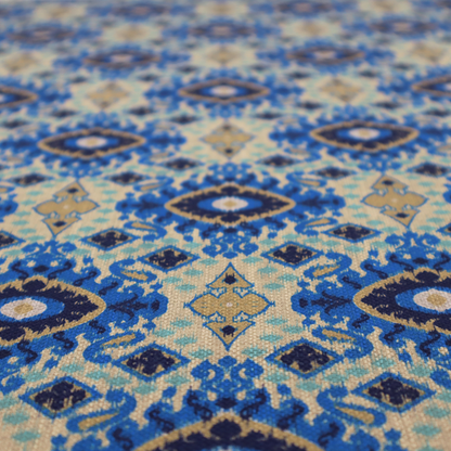 A repeating textile pattern featuring ornate blue Ikat medallions with a feathered, hand-painted appearance. The design includes smaller navy diamonds and sandy beige geometric shapes set against a light oatmeal-coloured fabric texture.