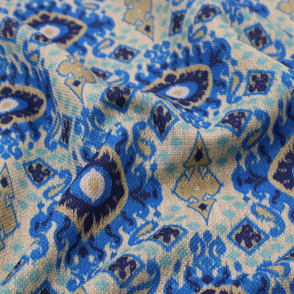 A repeating textile pattern featuring ornate blue Ikat medallions with a feathered, hand-painted appearance. The design includes smaller navy diamonds and sandy beige geometric shapes set against a light oatmeal-coloured fabric texture.