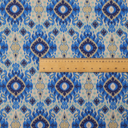 A repeating textile pattern featuring ornate blue Ikat medallions with a feathered, hand-painted appearance. The design includes smaller navy diamonds and sandy beige geometric shapes set against a light oatmeal-coloured fabric texture.