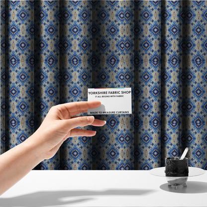 The fabric used to create these made-to-measure curtains is a repeating textile pattern featuring ornate blue Ikat medallions with a feathered, hand-painted appearance. The design includes smaller navy diamonds and sandy beige geometric shapes set against a light oatmeal-coloured fabric texture.