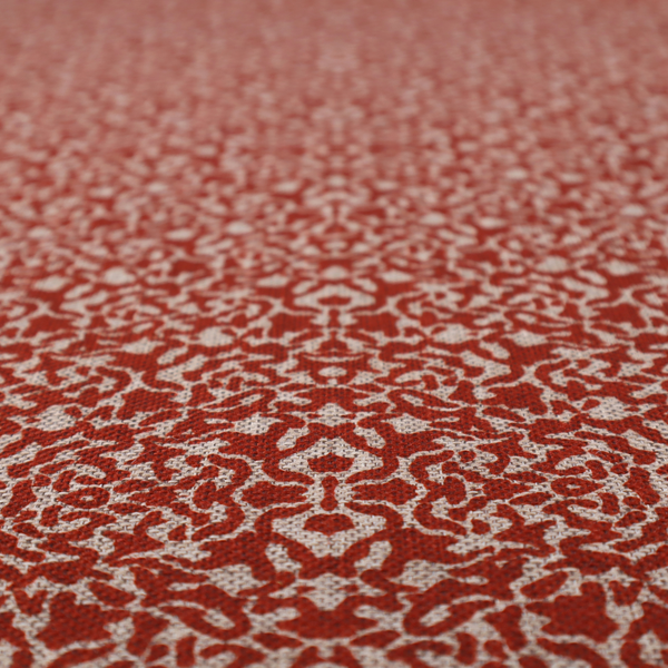 A decorative textile featuring a symmetrical, kaleidoscopic pattern in red and beige. The intricate, interlocking organic shapes create a dense, filigree-like design with a slightly weathered or distressed look. The fabric has a visible, fine-textured weave that gives the pattern a soft, matte appearance.