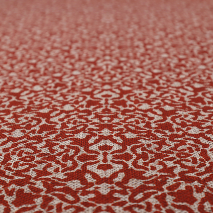 A decorative textile featuring a symmetrical, kaleidoscopic pattern in red and beige. The intricate, interlocking organic shapes create a dense, filigree-like design with a slightly weathered or distressed look. The fabric has a visible, fine-textured weave that gives the pattern a soft, matte appearance.