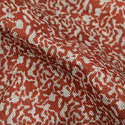 A decorative textile featuring a symmetrical, kaleidoscopic pattern in red and beige. The intricate, interlocking organic shapes create a dense, filigree-like design with a slightly weathered or distressed look. The fabric has a visible, fine-textured weave that gives the pattern a soft, matte appearance.