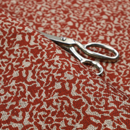 A decorative textile featuring a symmetrical, kaleidoscopic pattern in red and beige. The intricate, interlocking organic shapes create a dense, filigree-like design with a slightly weathered or distressed look. The fabric has a visible, fine-textured weave that gives the pattern a soft, matte appearance.