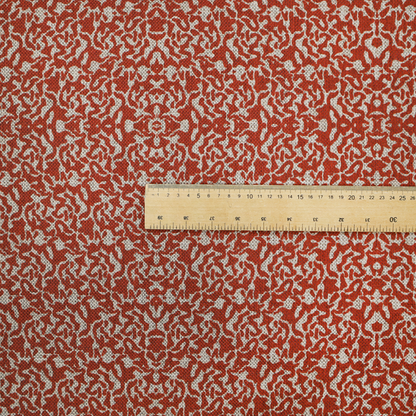 A decorative textile featuring a symmetrical, kaleidoscopic pattern in red and beige. The intricate, interlocking organic shapes create a dense, filigree-like design with a slightly weathered or distressed look. The fabric has a visible, fine-textured weave that gives the pattern a soft, matte appearance.