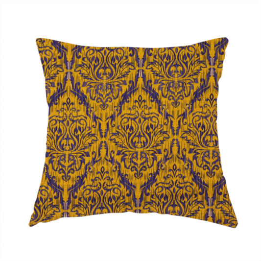 The fabric used to create this handmade cushion features a royal blue damask pattern layered over a deep mustard gold background. The design has an ikat-inspired finish, with vertical, feathered edges that give the ornate fleur-de-lis motifs a slightly blurred, textured appearance.