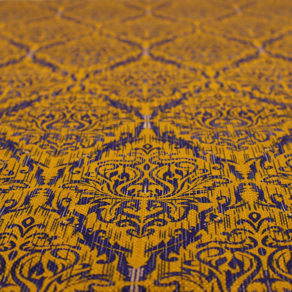 This fabric features a royal blue damask pattern layered over a deep mustard gold background. The design has an ikat-inspired finish, with vertical, feathered edges that give the ornate fleur-de-lis motifs a slightly blurred, textured appearance.