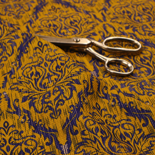 This fabric features a royal blue damask pattern layered over a deep mustard gold background. The design has an ikat-inspired finish, with vertical, feathered edges that give the ornate fleur-de-lis motifs a slightly blurred, textured appearance.
